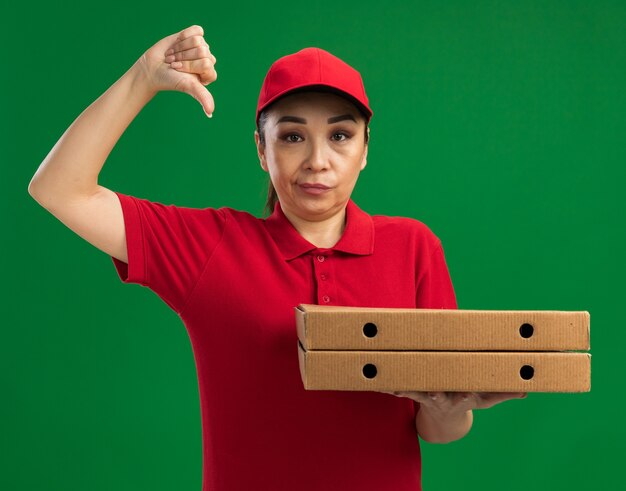 Young delivery woman in red uniform and cap holding pizza boxes  being displeased showing thumbs down standing over green wall