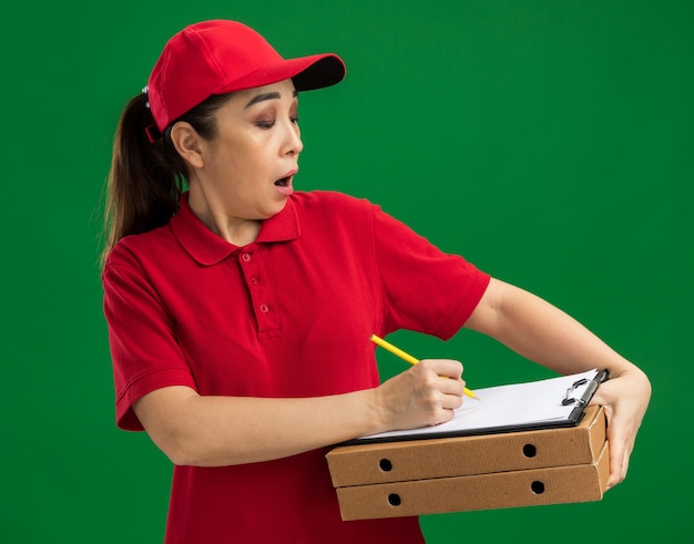 Young delivery woman in red uniform and cap holding pizza boxes and clipboard with pen writing something being surprised  