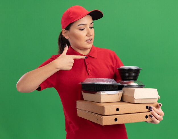緑の壁の上に立って混乱している人差し指で指しているピザの箱と食品パッケージを保持している赤い制服と帽子の若い配達の女性