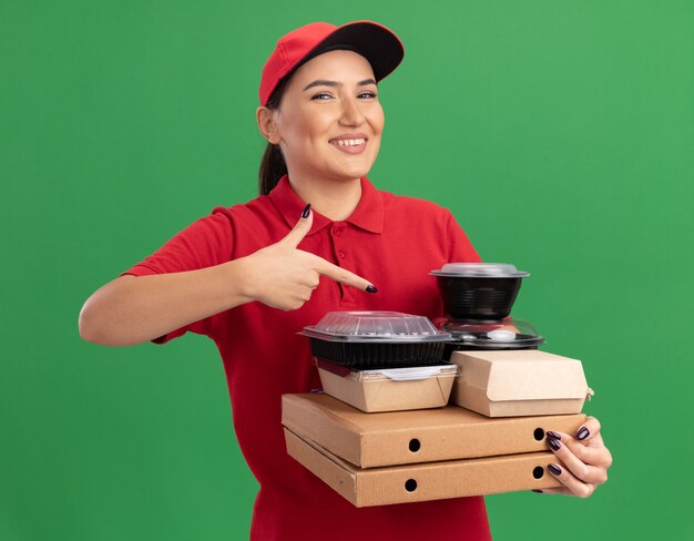 Young delivery woman in red uniform and cap holding pizza boxes and food packages pointing with index finger at them smiling cheerfully standing over green wall