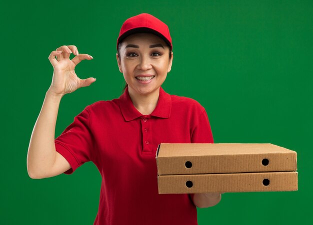 Giovane donna delle consegne in uniforme rossa e cappello che tiene in mano scatole per pizza che mostrano un gesto di piccole dimensioni che sorride con la faccia felice in piedi sul muro verde