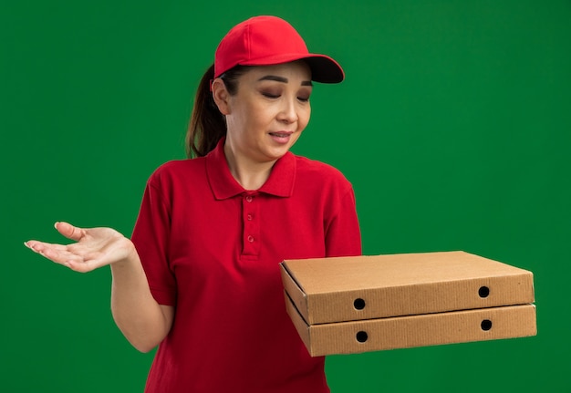 Молодая женщина-доставщик в красной форме и кепке держит коробки для пиццы, смущенные рукой, стоящей над зеленой стеной