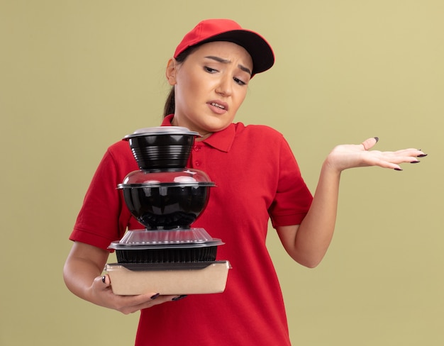 緑の壁の上に立っている答えがない混乱と不満に見える食品パッケージのスタックを保持している赤い制服と帽子の若い配達の女性