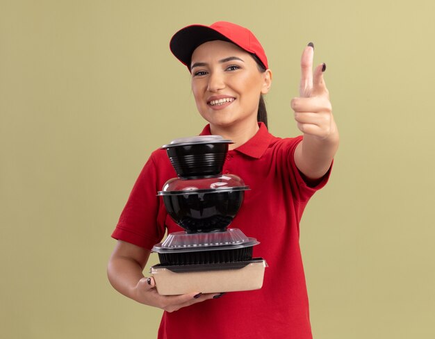 Foto gratuita giovane donna di consegna in uniforme rossa e cappuccio che tiene pila di confezioni di cibo che punta con il dito indice davanti sorridendo allegramente in piedi sopra la parete verde