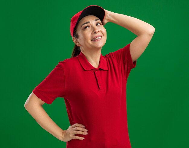 赤い制服を着た若い配達女性と、緑の壁の上に立っている彼女の頭に触れる混乱した表情で見上げる帽子