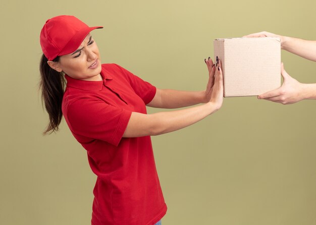 Young delivery woman in red uniform and cap refusing to take box package standing over green wall