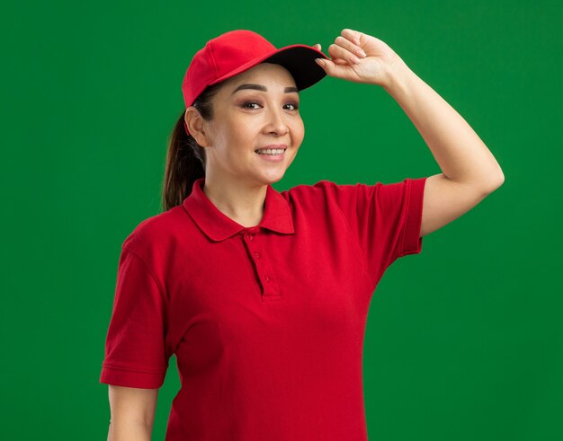 빨간 제복을 입은 젊은 배달 여자와 모자는 녹색 벽 위에 서있는 그녀의 모자를 만지고 자신감을 웃고