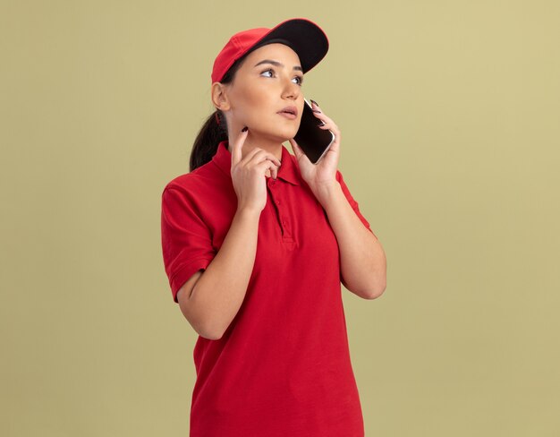 Молодая женщина-доставщик в красной форме и кепке разговаривает по мобильному телефону с серьезным лицом, стоящим над зеленой стеной