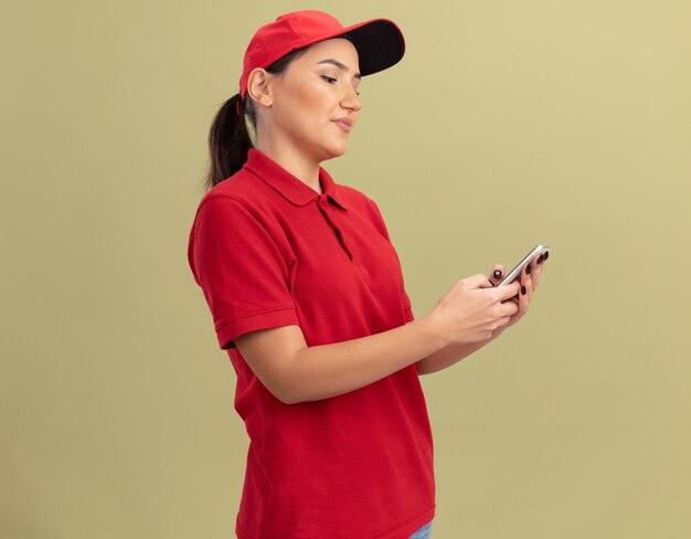 Young delivery woman in red uniform and cap using smartphone looking at it with smile on face standing over green wall