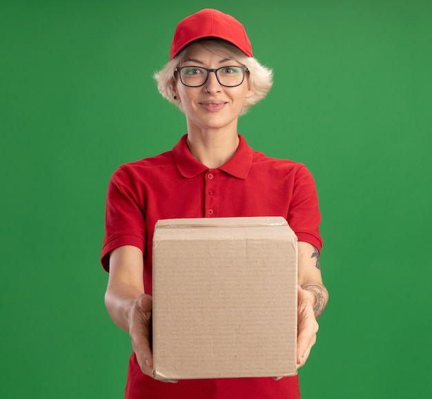 Young delivery woman in red uniform and cap wearing glasses holding cardboard box looking  smiling confident standing over green wall