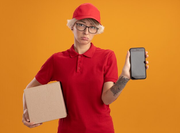 Young delivery woman in red uniform and cap wearing glasses holding cardboard box showing smartphone  with sad expression on face standing over orange wall