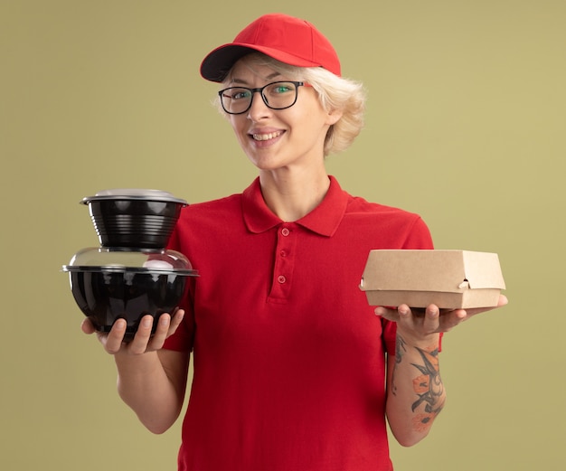 Молодая женщина-доставщик в красной форме и кепке в очках держит продуктовые пакеты, уверенно улыбаясь, стоя над зеленой стеной
