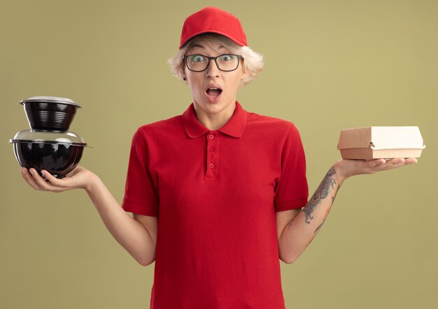 Young delivery woman in red uniform and cap wearing glasses holding food packages  surprised and amazed standing over green wall