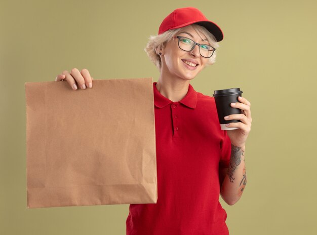 Young delivery woman in red uniform and cap wearing glasses holding paper package and coffee cup  smiling cherfully standing over green wall