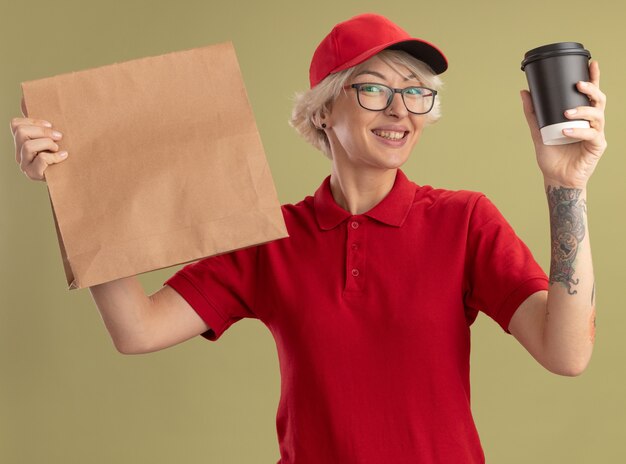 young delivery woman in red uniform and cap wearing glasses holding paper package and coffee cup  with happy face smiling standing over green wall