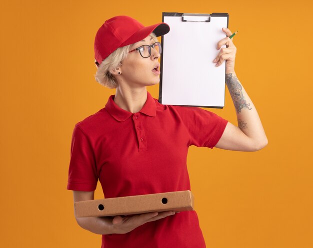 赤い制服を着た若い配達の女性とそれを見て興味をそそられるオレンジ色の壁の上に立って興味をそそられるピザボックスを示す眼鏡をかけている帽子