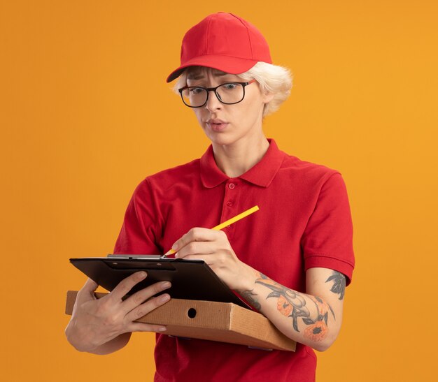 オレンジ色の壁の上に立って混乱しているように見えるクリップボードに何かを書いているピザボックスを保持している眼鏡をかけている赤い制服と帽子の若い配達の女性