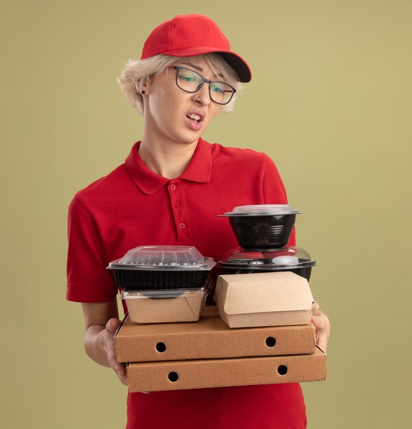 緑の壁の上に立っている悲しい表情でそれらを見ているピザボックスと食品パッケージを保持している眼鏡をかけている赤い制服と帽子をかぶった若い配達の女性