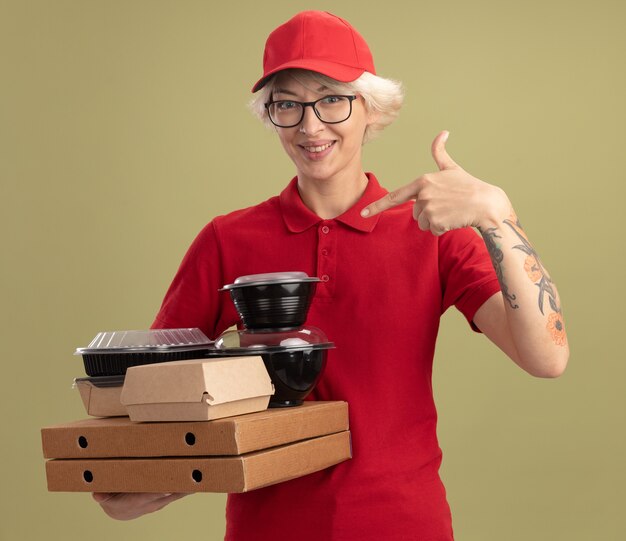 Giovane donna delle consegne in uniforme rossa e cappello con gli occhiali che tengono le scatole della pizza e i pacchetti di cibo che punta con il dito indice a loro sorridendo allegramente in piedi sopra la parete verde