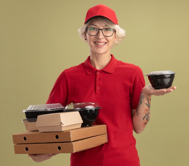 Young delivery woman in red uniform and cap wearing glasses holding pizza boxes and food packages  smiling with happy face standing over green wall