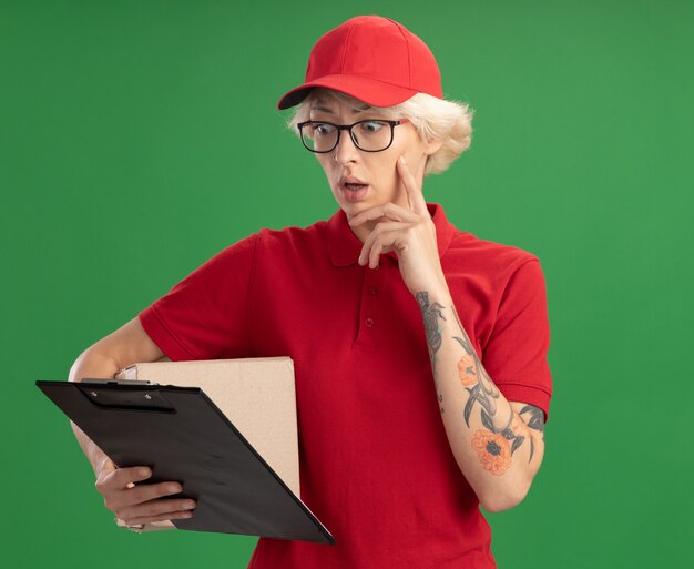 Молодая женщина-доставщик в красной форме и кепке в очках с картонной коробкой смотрит в буфер обмена в руке с смущенным выражением лица, стоя над зеленой стеной