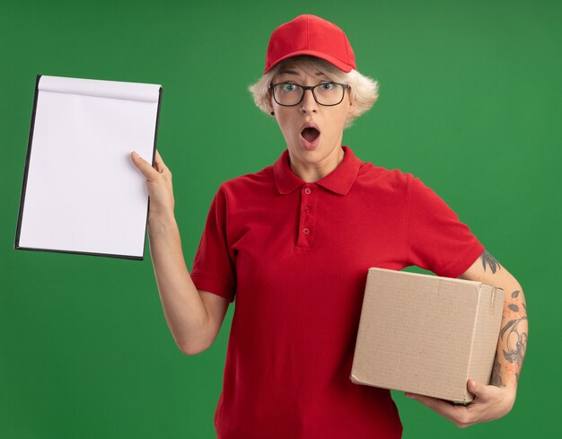 Young delivery woman in red uniform and cap wearing glasses with cardboard box showing clipboard with blank pages  worried and surprised standing over green wall