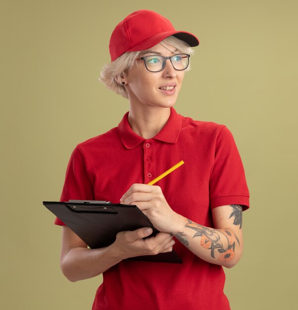 緑の壁の上に立っている顔に笑顔で脇を見てクリップボードと鉛筆で眼鏡をかけている赤い制服と帽子の若い配達の女性
