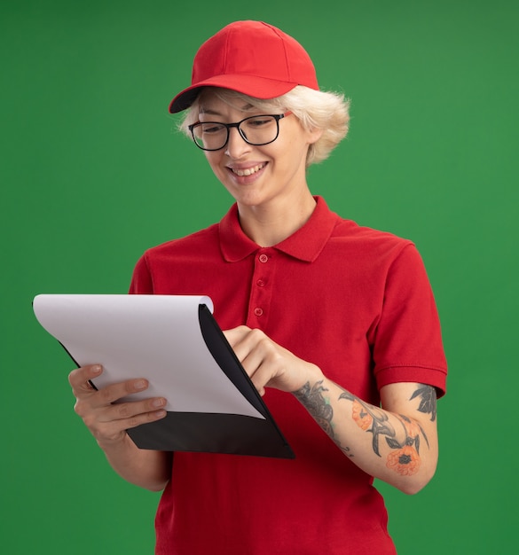 Young delivery woman in red uniform and cap wearing glasses with clipboard with blank pages looking at them smiling with happy face standing over green wall