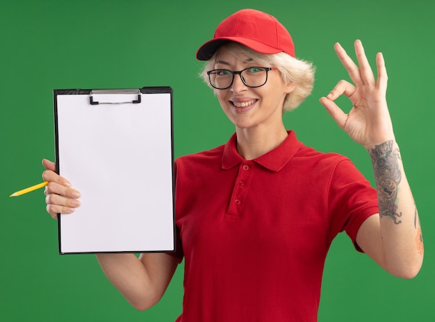 Young delivery woman in red uniform and cap wearing glasses with clipboard with blank pages and pencil  smiling cheerfully showing ok sign standing over green wall