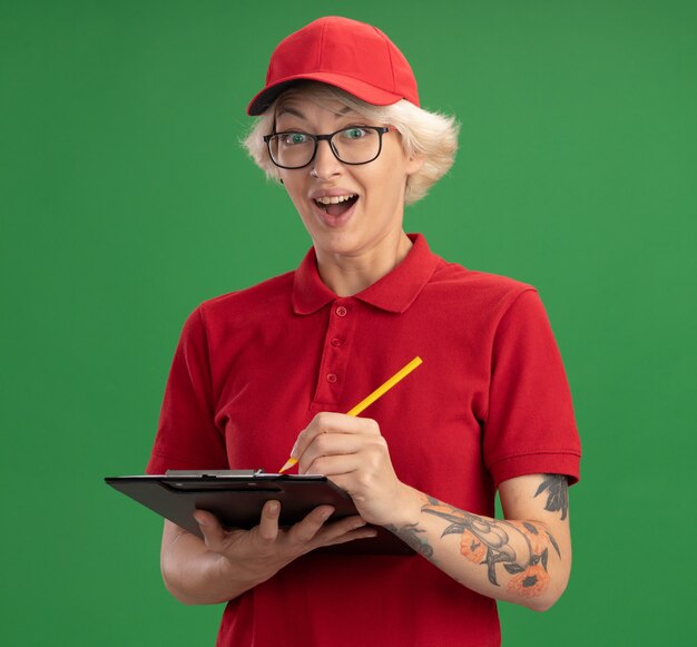 赤い制服とキャップを身に着けている若い配達の女性は、空白のページと鉛筆で眼鏡をかけて、緑の壁の上に立って幸せで驚きの笑顔を書いています