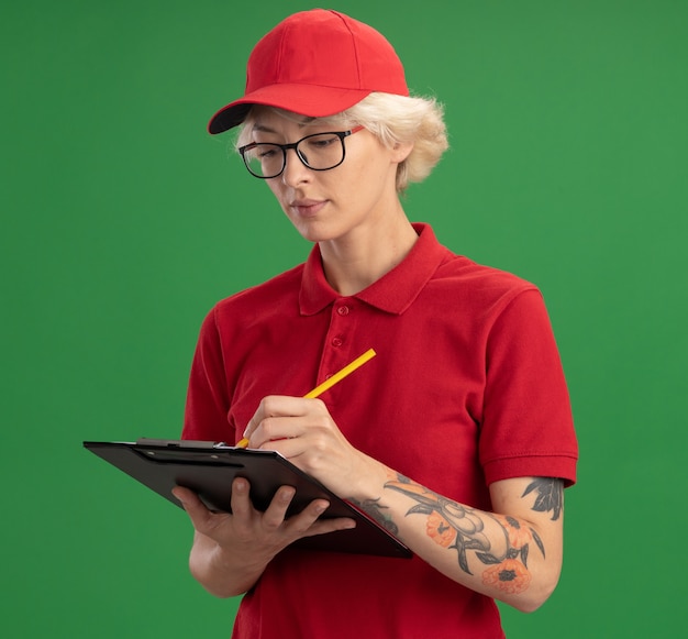 빨간색 유니폼과 모자에 젊은 배달 여자 빈 페이지와 녹색 벽 위에 서 심각한 얼굴로 뭔가 쓰는 연필 클립 보드와 안경을 쓰고