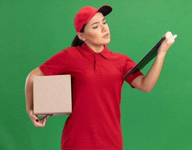 Young delivery woman in red uniform and cap with clipboard holding cardboard box looking at clipboard with serious face standing over green wall