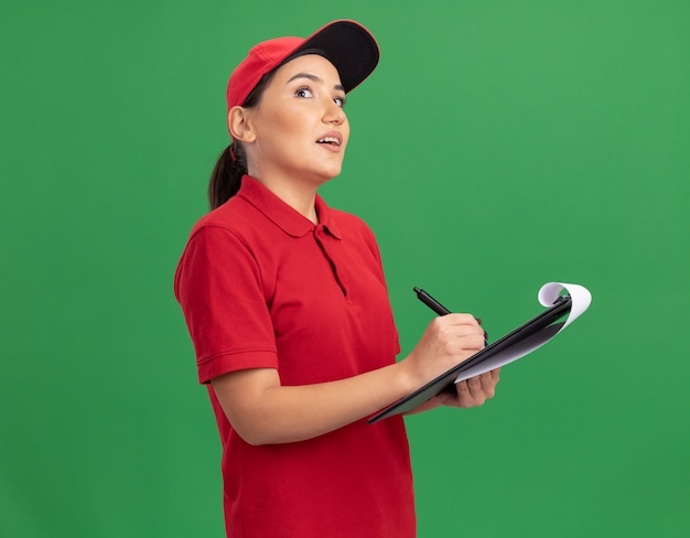 Young delivery woman in red uniform and cap with clipboard and pencil looking up with pensive expression thinking standing over green wall