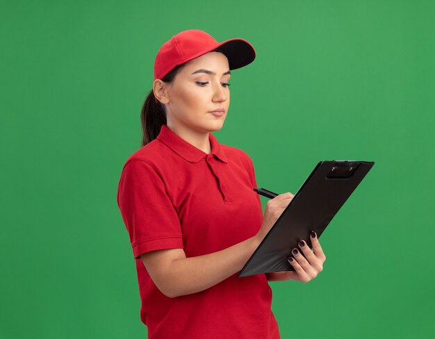 Young delivery woman in red uniform and cap with clipboard writing something with serious face standing over green wall