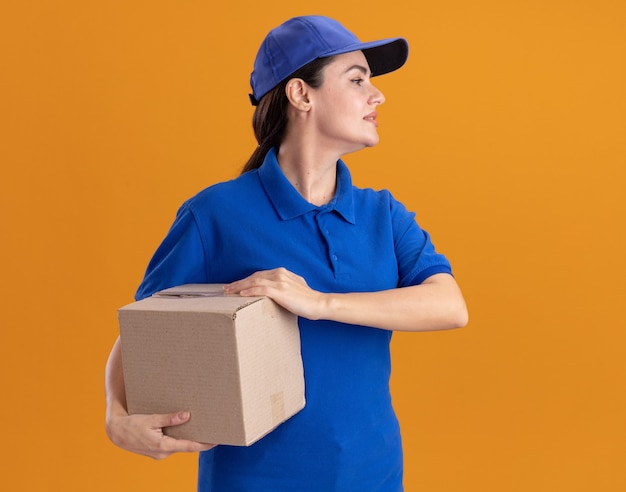 Молодая женщина-доставщик в униформе и кепке, держащая картонную коробку, смотрящую в сторону