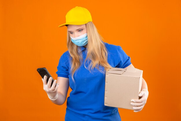 Young delivery woman wearing blue polo shirt and yellow cap in medical protective mask standing with cardboard box looking at screen of her smartphone over isolated dark yellow background