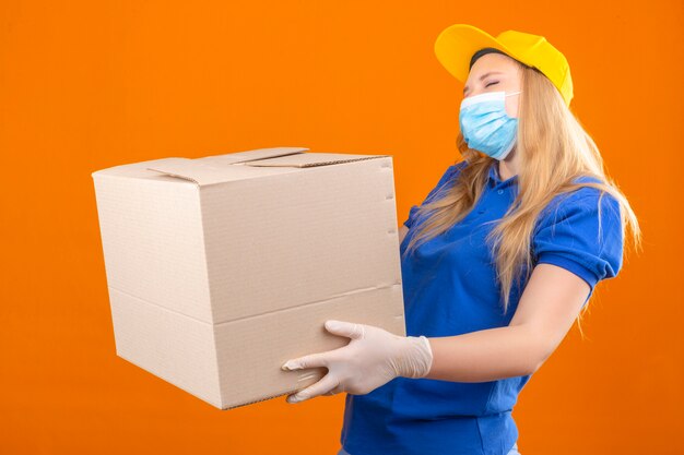 Young delivery woman wearing blue polo shirt and yellow cap in medical protective mask standing with cardboard box smiling cheerfully over isolated dark yellow background