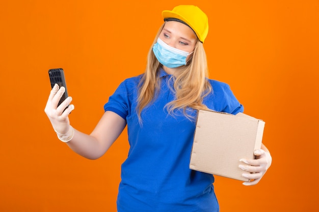 Young delivery woman wearing blue polo shirt and yellow cap in medical protective mask standing with cardboard box taking selfie on smartphone over isolated dark yellow background