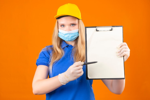 Young delivery woman wearing blue polo shirt and yellow cap in medical protective mask standing with clipboard pointing with pen asking for signature with serious face over isolated dark yellow