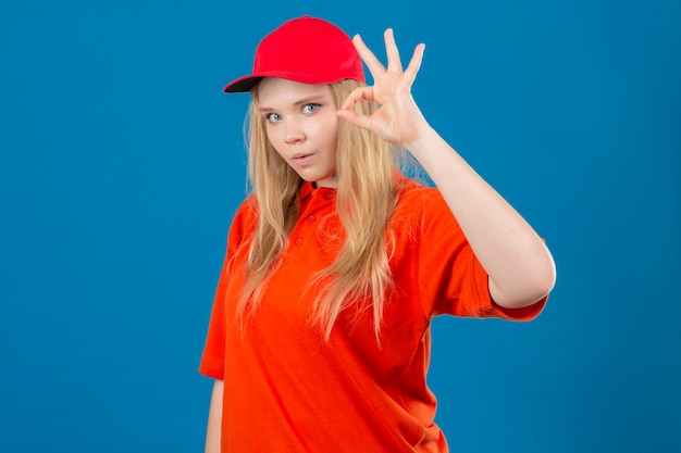 오렌지 폴로 셔츠와 빨간 모자를 쓰고 젊은 배달 여자는 고립 된 파란색 배경 위에 확인 서명을하고 놀란 찾고