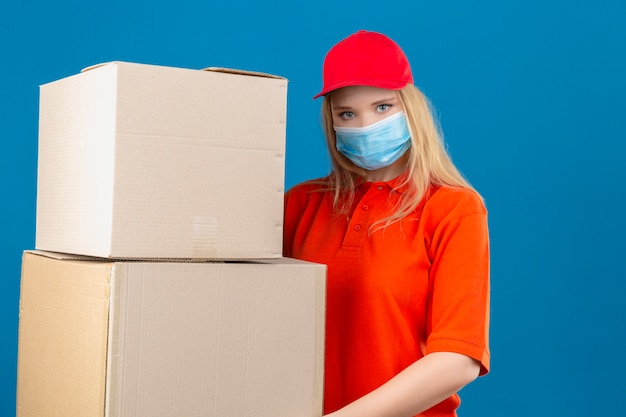 Young delivery woman wearing orange polo shirt and red cap in medical protective mask holding large cardboard boxes looking at camera with serious face over isolated blue background