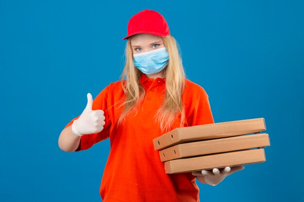 Young delivery woman wearing orange polo shirt and red cap in medical protective mask holding a stack of pizza boxes showing thumb up smiling over isolated blue background