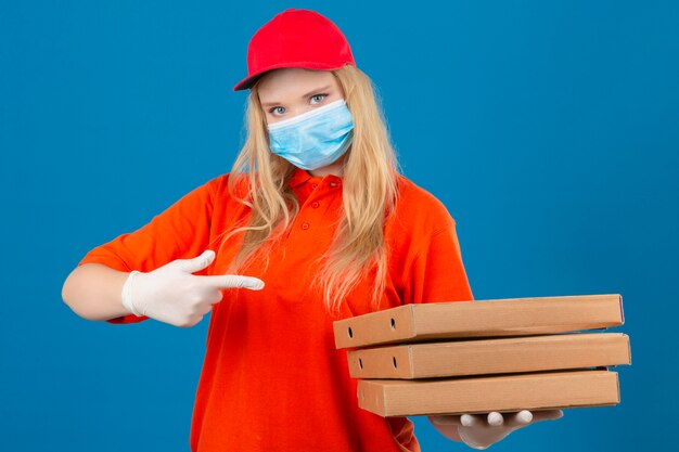 Молодая женщина-доставщик в оранжевой рубашке поло и красной кепке в медицинской защитной маске, указывая на стопку коробок из-под пиццы в другой руке, уверенно смотрит на изолированный синий фон