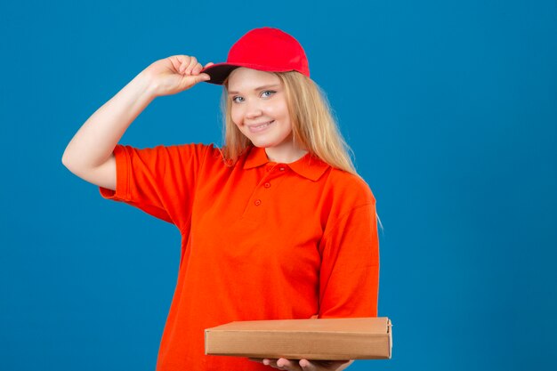 Молодая курьерская женщина в оранжевой рубашке поло и красной кепке приветствует улыбку и дружелюбно трогает ее кепку, стоящую на изолированном синем фоне