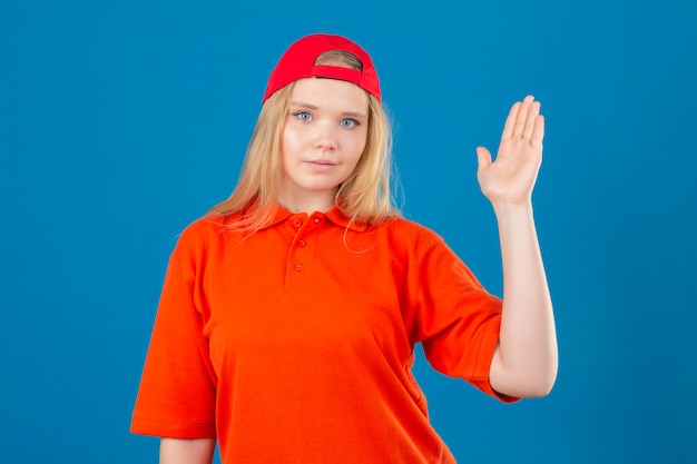 Young delivery woman wearing orange polo shirt and red cap smiling friendly waving hand welcoming and greeting you or saying goodbye over isolated blue background