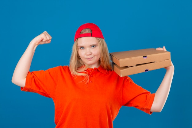 Free photo young delivery woman wearing orange polo shirt and red cap standing with pizza boxes on shoulder raising fist like a winner over isolated blue background