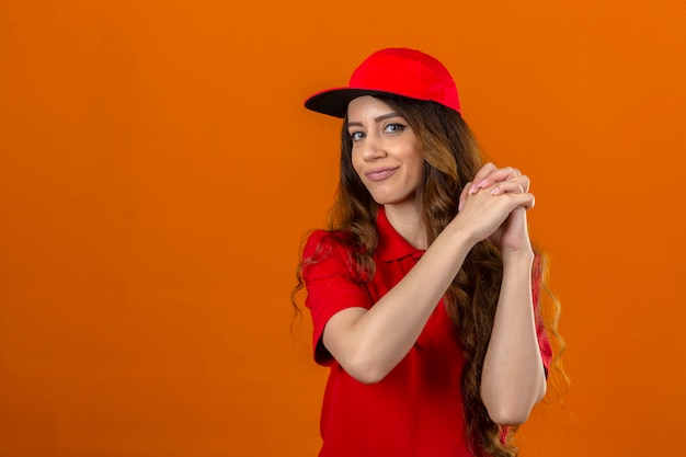 Молодая доставщица в красной рубашке поло и кепке жестикулирует со сложенными руками, выглядит уверенно и гордо на изолированном оранжевом фоне