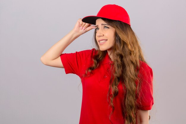 Young delivery woman wearing red polo shirt and cap having doubts while touching head over isolated white background