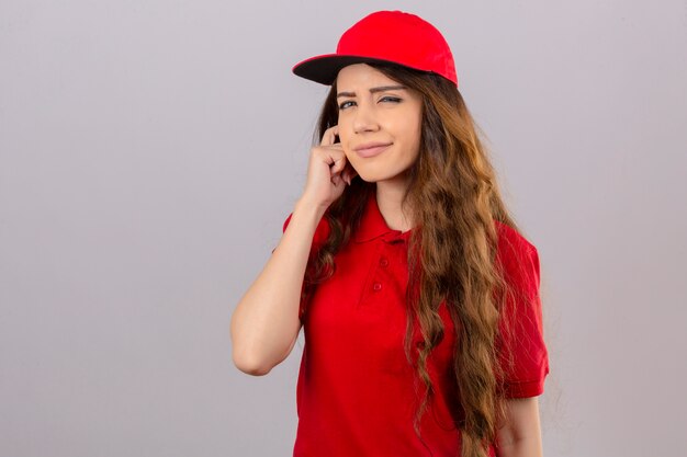 Young delivery woman wearing red polo shirt and cap looking at camera squinting looking at camera with suspicion over isolated white background