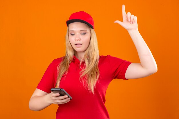 Foto gratuita giovane donna delle consegne indossando maglietta polo rossa e cappuccio guardando il telefono cellulare in mano e puntare il dito indice verso l'alto avente un nuovo concetto di idea isolato su sfondo arancione